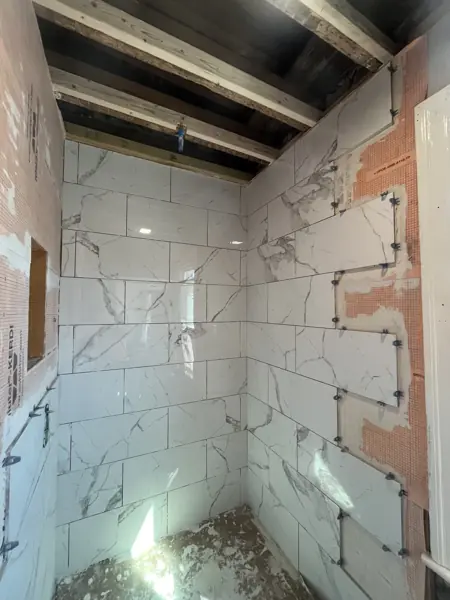 Large white marble-look tiles being set on the shower walls with leveling clips, with orange membrane visible where tile is not yet installed.
